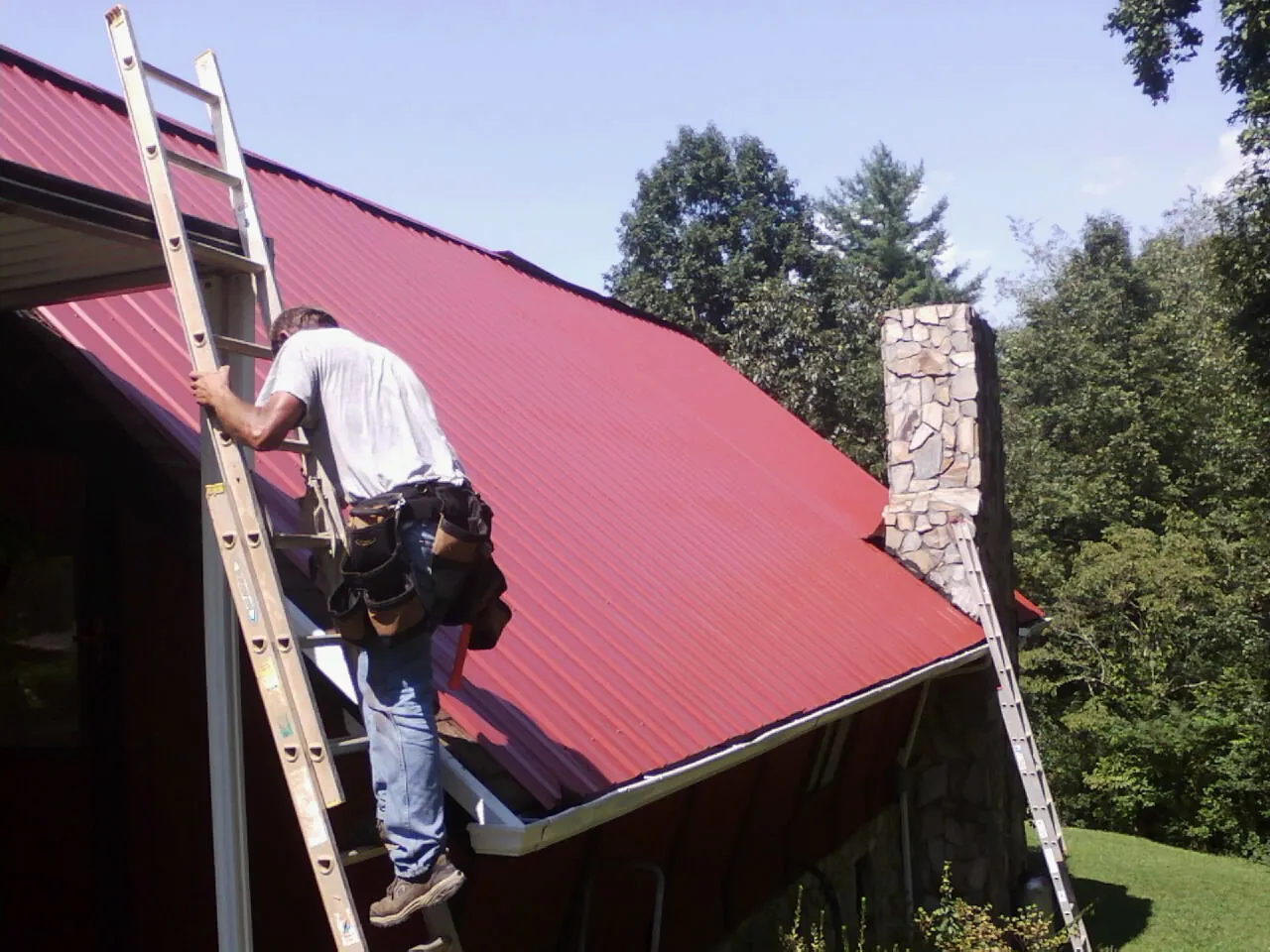 Roofer on a ladder beside a red metal roof and stone chimney — Soffit & Fascia Repair by Roofing Brothers in York Springs